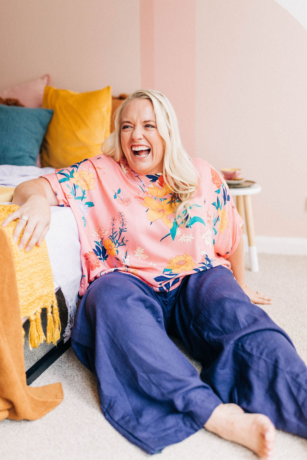 woman laughing, leaning against a bed and wearing a pink floral adaptive top and navy linen adaptive pants 