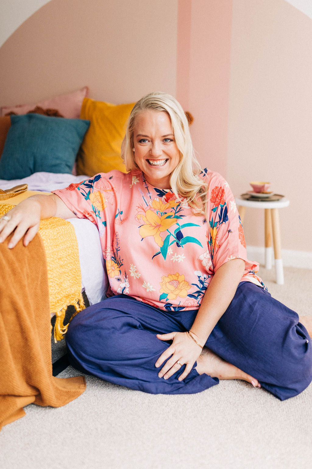 smiling woman sitting comfortably on the floor wearing a pink floral adaptive top and adaptive linen pants in navy