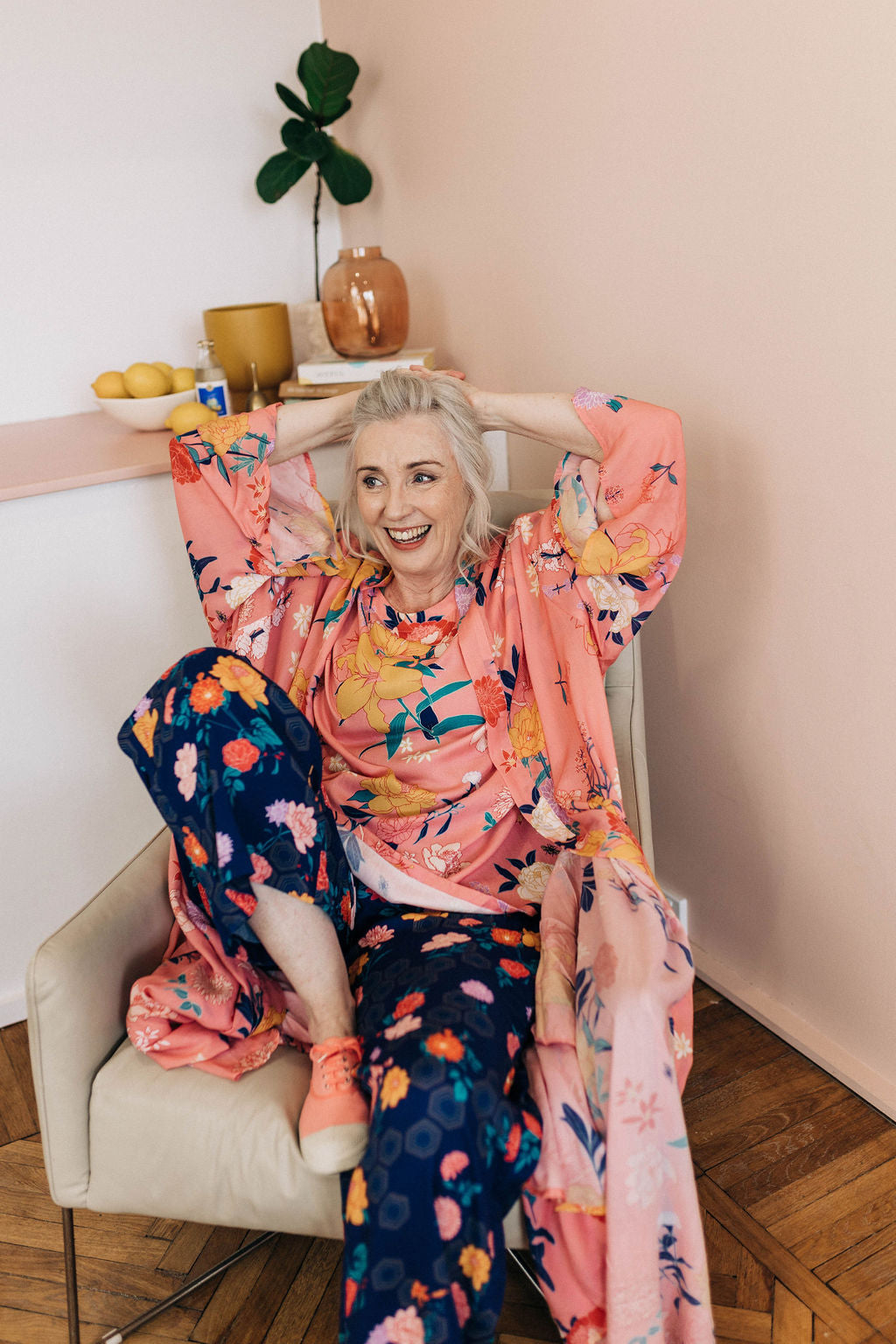 woman with navy floral geometric print adaptive pants and matching limonata pink floral top and robe smiling with her arms over her head on a cream leather chairand robe